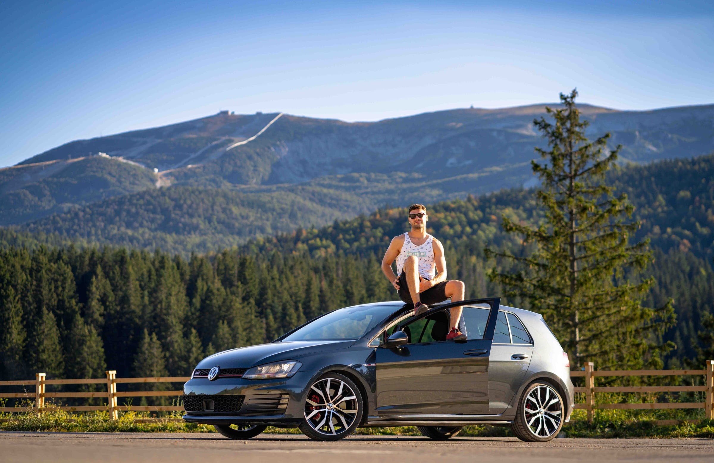 A grey Volkswagen Golf with a man sitting on its roof, surrounded by forest and mountains.